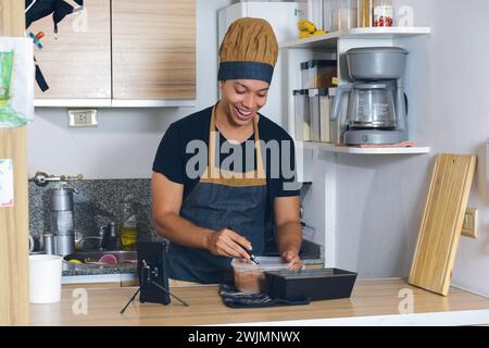 Junger lateinamerikanischer Mann in Küchenuniform zu Hause, glücklich, Kuchen für seine Familie zuzubereiten und Video-Tutorial mit Telefon zu sehen. Stockfoto
