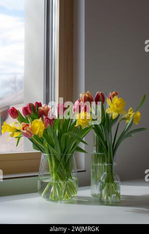 Glasvasen mit Frühlingsblumen auf einem weißen Tisch in der Nähe des Fensters Stockfoto