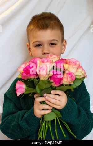 Ein kleiner Junge in einem grünen Pullover gibt einen Strauß rosa Rosen Stockfoto