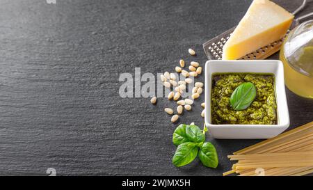 Hausgemachte Pesto-Sauce im kleinen quadratischen Glas und Zutaten für Pasta auf schwarzem Schieferhintergrund mit Kopierraum. Traditionelle italienische Küche, Rezept, Res Stockfoto