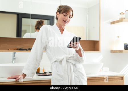 Kaukasische Frau mittleren Alters in einem weißen Gewand genießt ihr Telefon zu Hause Stockfoto