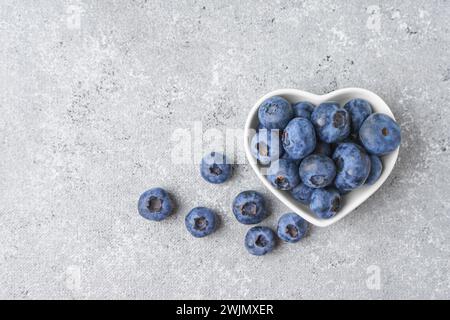 Frische Heidelbeeren und Verstreuen von Beeren in kleiner weißer herzförmiger Schüssel auf grauem Betonhintergrund. Bio-Beeren, gesundes Essen, wilde Beeren. Stockfoto