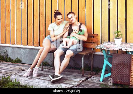 Junge Eltern von 26 Jahren mit einem sechsmonatigen Kind in den Armen sitzen im Sommer im Hinterhof ihres Hauses. Stockfoto