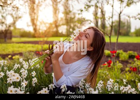 Porträt junger Gärtner, die Narzissen im Frühlingsgarten pflücken. Glückliche Frau, die Narzissen riecht, Blüten, geschnitten mit Gartenschere bei Sonnenuntergang. Gartenarbeit Stockfoto