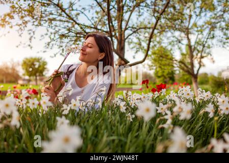 Junger lächelnder Gärtner pflückt Narzissen im Frühlingsgarten. Glückliche Frau, die Narzissen riecht, Blüten, geschnitten mit Gartenschere bei Sonnenuntergang. Gartenarbeit Stockfoto