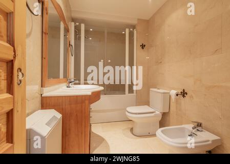 Badezimmer, cremefarbene Marmorfliesen, Kirschholzschrank mit integriertem Spiegel und Waschbecken mit Marmorarbeitsfläche, weiße Toiletten und Duschkabine mit Glas Stockfoto