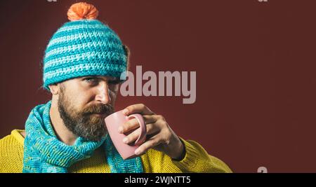 Ernster, gutaussehender Mann mit Bart und Schnurrbart in gelbem Pullover, blauem Schal und Hut mit einer Tasse Kaffee oder Tee. Stilvoller bärtiger Mann, der heiß trinkt Stockfoto