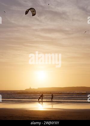 Kitesurfer an einem Sandstrand bei Sonnenuntergang mit einer Insel dahinter, während die Wellen hereinrollen, in Essaouira, Marokko. Februar 2024 Stockfoto