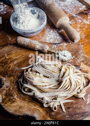 Nahaufnahme von handgemachter Pasta namens Fettuccine, die auf einem hölzernen Schneidebrett ruhen gelassen wurde. Einige nützliche Werkzeuge: Einen Spannstift und einen Teigschneider. Stockfoto