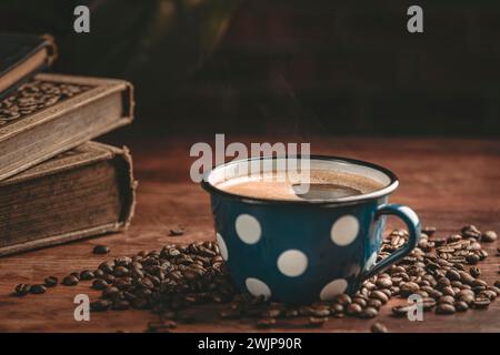 Dampfend blaue Kaffeetasse umgeben von Kaffeebohnen und alten Büchern auf einem Holztisch Stockfoto