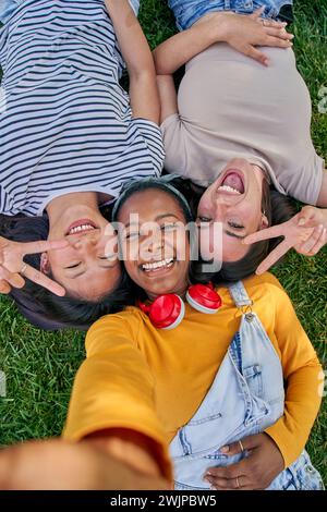 Vertikales Selfie über drei fröhlichen, multirassischen jungen Frauen, die draußen auf Gras liegen. Gen z in Parkstellung. Stockfoto