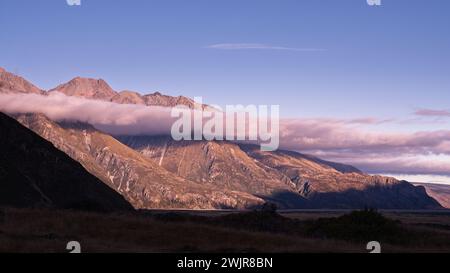 Herrliches Glühen bei Sonnenuntergang: Der Berggipfel erstrahlt in warmem Licht inmitten atemberaubender lila Wolken. Stockfoto