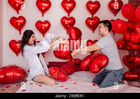 Verspielter Moment zwischen Paaren, die sich in Kissenballons auf dem Bett kämpfen, mit herzförmigen roten Ballons im Hintergrund. Die Frau macht fröhlich rumtäuscht Stockfoto