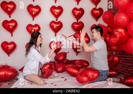 Verspielter Moment zwischen Paaren, die sich in Kissenballons auf dem Bett kämpfen, mit herzförmigen roten Ballons im Hintergrund. Die Frau macht fröhlich rumtäuscht Stockfoto