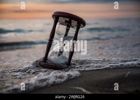 Sanduhr am Ufer mit Wellen, die die flüchtige Natur der Zeit bei Sonnenuntergang widerspiegeln. Stockfoto