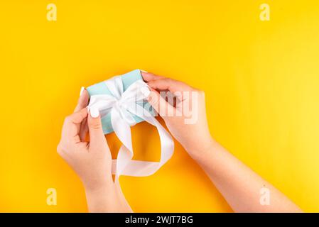 Foto von der ersten Person von oben mit weiblichen Händen, die eine blaue Geschenkbox mit weißem Band auf gelbem Hintergrund halten. Meine Hände wiegen eine pastellblaue Geschenkbox Stockfoto