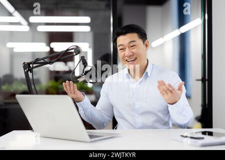 Ein begeisterter asiatischer Geschäftsmann nimmt einen Podcast in einer modernen Büroumgebung auf, der Glück und Professionalität zum Ausdruck bringt. Stockfoto