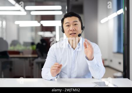 Asiatischer Geschäftsmann mit Headset im modernen Büro, der Kundensupport bietet oder ein Team-Meeting leitet und emotionale Bindung zeigt. Stockfoto
