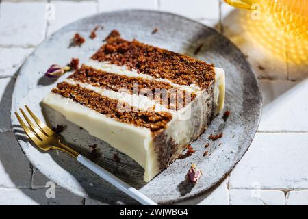 Ein Stück Karottenkuchen, serviert auf weißem Fliesentisch. Helle, sonnige Szene. Lagenkuchen mit Frischkäse-Zuckerguss und flauschigem Biskuit mit Getränk Stockfoto