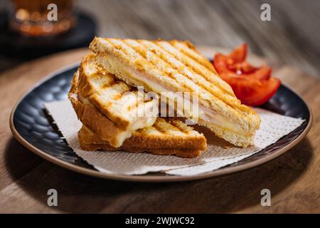 Geschnittenes Toast-Sandwich mit geschmolzenem Gouda-Käse und putenschinken auf hölzernem Hintergrund mit Kirschtomaten auf einem schwarzen Teller. Serviert mit Eistee. Stockfoto