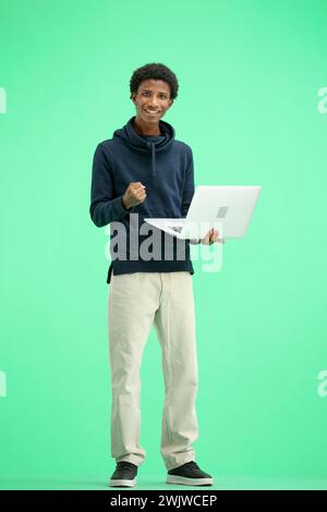 Ein Mann in voller Länge, auf grünem Hintergrund, mit einem Laptop, zeigt die Daumen hoch Stockfoto