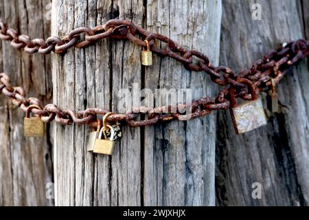 Verwitterte Holzstange, umwickelt mit einer rostigen Kette und drei verschiedenen Vorhängeschlössern, symbolisiert dauerhafte Liebe und Engagement Stockfoto