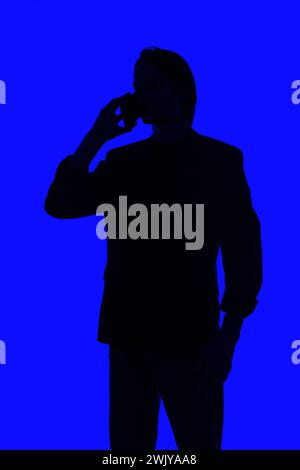 Unerkennbare Silhouette ein eleganter Geschäftsmann, der auf einem Smartphone spricht, in einer Jacke, die auf einem blau leuchtenden Hintergrund steht, ein futuristisches Konzept Stockfoto