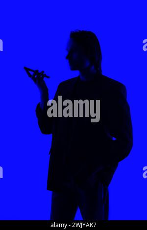 Silhouette ein eleganter Geschäftsmann, der auf einem Mobiltelefon spricht, in einer Jacke, die auf einem blau leuchtenden Hintergrund steht, futuristische KI-Konzepte Stockfoto