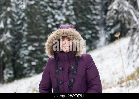 Schöne elegante, attraktive glückliche ältere Frau mittleren Alters im Schnee mit schneebedeckten Bäumen, die eine lila Jacke und eine Pelzhaube tragen. Stockfoto