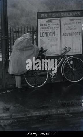 Eine Radfahrerin in einem wasserdichten cape justiert ihr Fahrrad 1950 vor einem britischen Bahnhof. Dieses Bild ist ein Foto, das vom ursprünglichen negativ aufgenommen wurde. Es könnte zu Unreinheiten führen. Es gibt eine Hinweistafel, in der Bahnfahrten nach London und Southport angekündigt werden. Stockfoto