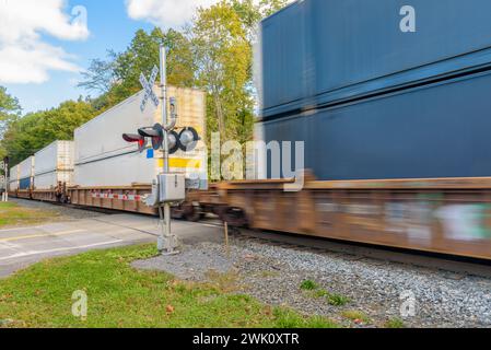 Schnell fahrender Containerzug, der an einem sonnigen Herbstmorgen an einer Eisenbahnüberquerung vorbeifährt Stockfoto