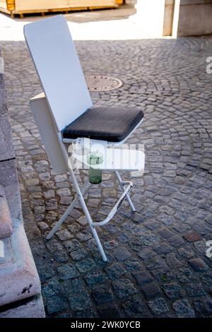 Auf Kopfsteinpflaster steht ein weißer Klappstuhl aus Holz und Metall mit einem kleinen anhängenden Tisch mit einem Getränkehalter. Es ist ein schwarzes Kissen auf Stockfoto