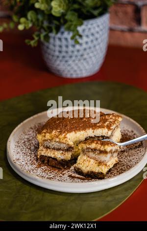 Nahaufnahme einer Portion Tiramisu-Kuchen mit Kakaopulver, serviert mit einer Gabel auf einem weißen Teller. Grüne Pflanze in einer Vase auf dem Restauranttisch, b Stockfoto