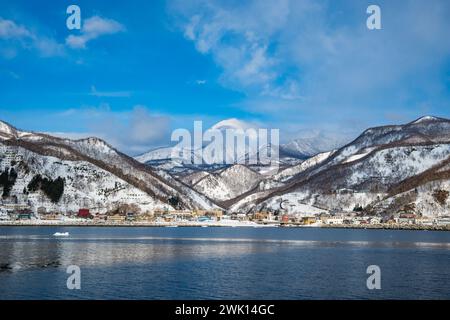 Kleine Stadt an der Küste, mit schneebedeckten Bergen im Hinterland. Rausu, Hokkaido, Japan. Stockfoto
