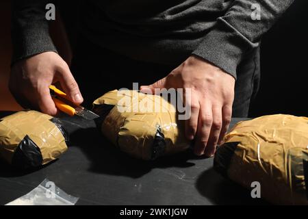 Schmuggel und Drogenhandel. Mann, der ein Paket mit Betäubungsmitteln öffnet, mit einem Cutter am dunklen Tisch, Nahaufnahme Stockfoto