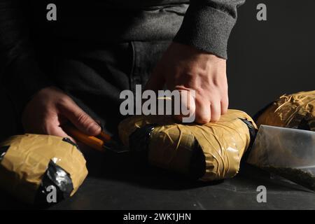 Schmuggel und Drogenhandel. Mann, der ein Paket mit Betäubungsmitteln öffnet, mit einem Cutter am dunklen Tisch, Nahaufnahme Stockfoto