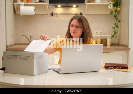 Eine Frau mit einem Laptop druckt Papier auf einem Drucker, während sie an einem Tisch in der Küche sitzt. Eine erwachsene Geschäftsfrau arbeitet von zu Hause aus Stockfoto