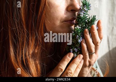 Rothaarige Frau, die einen Haufen blühenden Rosmarin in den Händen hält und seinen Duft einatmet. Kräuterkundige Frau, die frische Bio-Kräuter für Naturkraut zubereitet Stockfoto