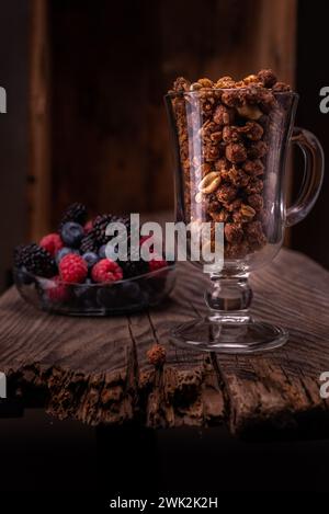 Schokoladengranola in einem Glas. Bunte Beeren im Hintergrund. Frühstück mit Müsli und Beeren. Vertikaler Rahmen. Stockfoto