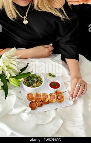 Raffinierte Frau, die sich auf weißer Bettwäsche mit Tablett mit köstlichen frisch gebackenen Würstchen, gesundem Gemüsesalat, scharfen Dippsoßen und f Stockfoto