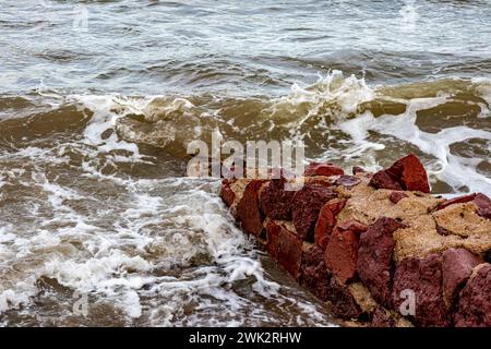 Kleine rötlich-braune unregelmäßige Steinmauer am Strand, Meereswellen brechen an der felsigen Küste, bilden reichlich Schaum, unebene Oberfläche, bewölkter Tag Stockfoto