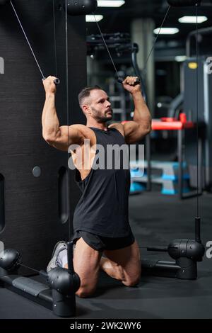 Ernsthafter Sportler mit schwarzem Tanktop, Sport für Bizeps, auf Knien. Seitenansicht eines muskulösen jungen Mannes, der an den Kabeln des Trainingsapparates zieht und die Muskeln in Spannung hält. Sportkonzept. Stockfoto