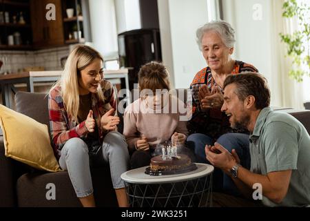 Eine herzerwärmende Szene entfaltet sich, während sich eine Familie mit mehreren Generationen auf einer Couch versammelt, um einer entzückten Großmutter einen Geburtstagskuchen zu präsentieren und Erinnerungen zu schaffen Stockfoto