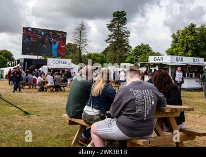 Beim Goodwood Festival von Speed West Sussex UK saßen die Leute um Bänke mit Lieferwagen und servierten Speisen und Getränke und schauten sich eine Großleinwand an Stockfoto