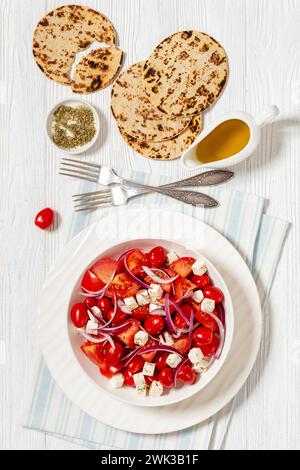 Tomatensalat mit roten Zwiebeln und weißem salzigem griechischen Käse in Schüssel auf weißem strukturiertem Holztisch mit Gabeln und flachen Brotkreisen, vertikale Ansicht von Stockfoto