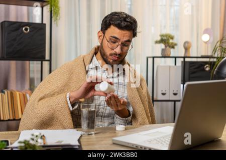 Gesundheit, Kälte und Menschen Konzepte. Der kranke junge hispanische Geschäftsmann, in eine Decke gehüllt, arbeitet am Laptop am Schreibtisch im Heimbüro. Ungesunder Hispanic Freelancer nimmt Medikamente und zittert vor der Kälte zu Hause Stockfoto