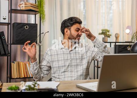 Überarbeiteter junger indischer arabischer Geschäftsmann, der unter Augenanstrengung und Kopfschmerzen leidet und am Laptop arbeitet, der am Schreibtisch im Heimbüro sitzt. Müde Freelancer zieht Brillen und Massagen aus, reibt Nasenrücken. Stockfoto