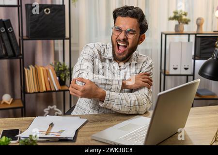 Müder indischer arabischer Geschäftsmann Guy Manager, der mit Laptop am Tisch sitzt und Schmerzen und Beschwerden im Ellenbogengelenk hat und schmerzhafte Hände massiert. Arthritis- und Bursitis-Konzepte Stockfoto