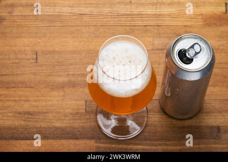 Kaltes Bierglas gefüllt mit einem goldenen Bier, gekrönt mit einem cremigen Kopf neben einer offenen Bierdose mit Kondenswasser auf einem natürlichen Holztisch Stockfoto