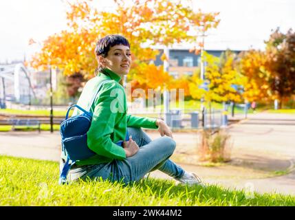 Lächelnde junge Frau in grünem Pullover mit blauem Rucksack, die an einem sonnigen Herbsttag im Park auf dem Gras sitzt. Unbeschwerte Dame entspannt ihn Stockfoto